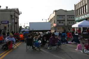 Outdoor movie setup + group of people sitting in lawn chairs near white semi truck