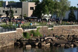 Large group of people standing and sitting along side of river listening to live music