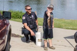 Police officer kneeling next to kid holding fish near river